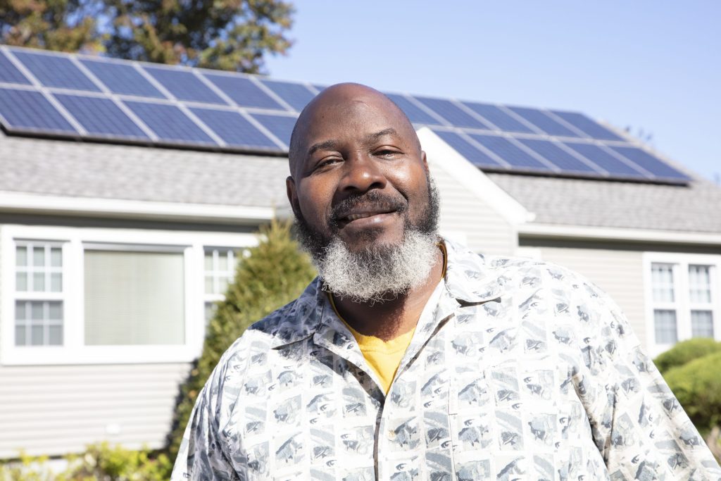 Peter smiling outside their home, with solar panels on the roof behind